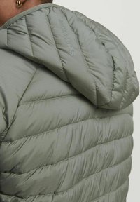 Grüne Pufferjacke mit gepolstertem Design, strukturiertem Obermaterial und Kapuze. Der Markenname "JACK & JONES" ist auf der Schulter eingraviert.