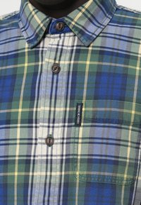 Chemise en flanelle à carreaux avec des motifs en plaid verts, bleus et jaunes, avec une fermeture par bouton et une poche poitrine avec une étiquette de marque.