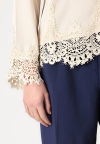 Top beige con raffinato bordo in pizzo crema, caratterizzato da motivi dettagliati e una consistenza morbida, abbinato a pantaloni blu navy scuri.