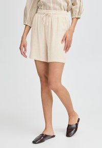 Pantaloni corti beige con vita elastica e cordoncino, realizzati in tessuto testurizzato. Indossati con scarpe scivolose marrone scuro.