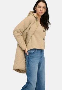 Beige kviltet lang frakke med hætte, parret med en lys tan tekstureret sweater med knapdetaljer og blå denim jeans.
