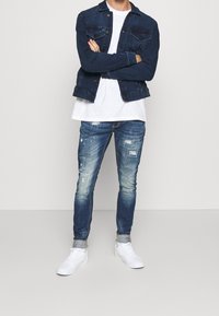 Tummansininen farkkutakki, jossa on nappikiinnitys ja rintataskut. Alla valkoinen T-paita. Haalistuneet siniset slim fit -farkut, joissa on kulutettuja yksityiskohtia. Valkoiset sneakerit.