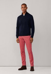 Tengerészkék cipzáras nyakú pulóver, piros chino nadrág és sötétbarna csizma. A pulóver puha textúrájú, a nadrág pedig egyenes szárú.