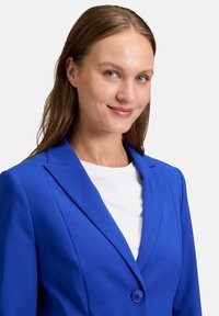 Modrý blazer s klopami a zapínáním na jeden knoflík, vyrobený z hladkého materiálu, doplněný bílým topem pod ním.