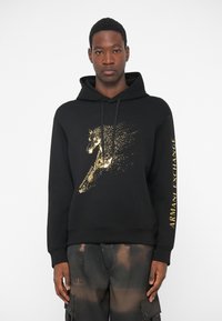 Μαύρη φόρμα με κουκούλα που διαθέτει ένα χρυσό γραφικό άλογο και κείμενο "ARMANI EXCHANGE" στο μανίκι. Μαλακό υλικό, κανονική εφαρμογή και κουκούλα με κορδόνια.