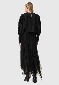 Γυναίκα με την πλάτη στραμμένη, φοράει μαύρο μπλούζα με μακριά μανίκια και λεπτομέρειες από δαντέλα, καθώς και μακριά φούστα από δαντέλα που ταιριάζει, με μποτάκια μέχρι τον αστράγαλο.