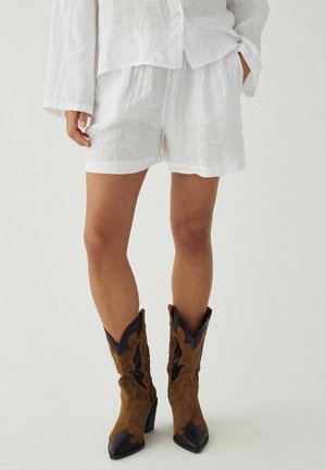 Fehér lenvászon rövidnadrágot és gombos inget viselő személy, barna bőr western stílusú félcsizmával, egyszínű háttér előtt.