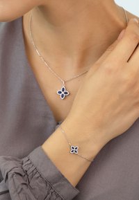 Silberne Halskette und Armband mit einem Blumendesign, versehen mit dunkelblauen Steinen, auf einer strukturierten blauen Bluse.