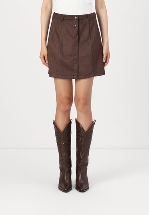 Jupe en suède marron avec boutons devant, poches et coupe ajustée. Portée avec des bottes de cow-boy marron montant jusqu'aux genoux, à bout pointu.