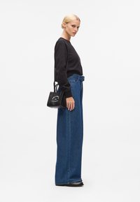 Svart langermet skjorte, brede blå denimjeans og liten svart veske med hvit tekst. Modell står i profil mot en hvit bakgrunn.