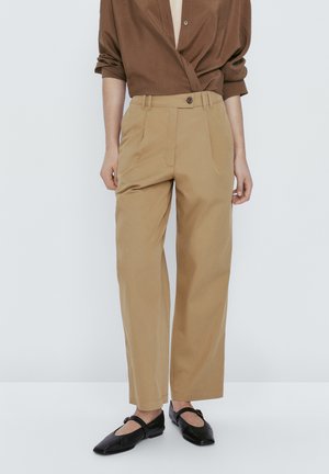 Person trägt beige hochtaillierte, gerade geschnittene Hose, braunes Langarmshirt mit aufgekrempelten Ärmeln und schwarze Mary-Jane-Ballerinas.
