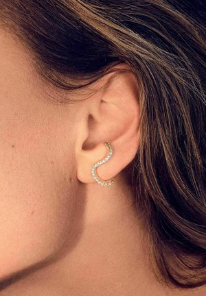 Guld bøjet ørestik med bølgemønster, prydet med små klare krystaller, vist i et øre imod en blød baggrund.