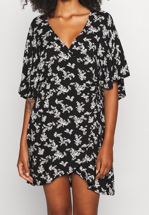 Zwarte wikkeljurk met witte bloemenpatronen, V-hals, korte fluttermouwen en asymmetrische zoom, gedragen door een persoon met een getatoeëerde arm.