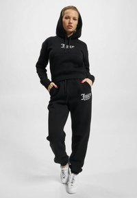 Černá croppovaná mikina s kapucí a přední kapsou, doplněná o černé tepláky s detailním logem. Model má na sobě bílé tenisky.