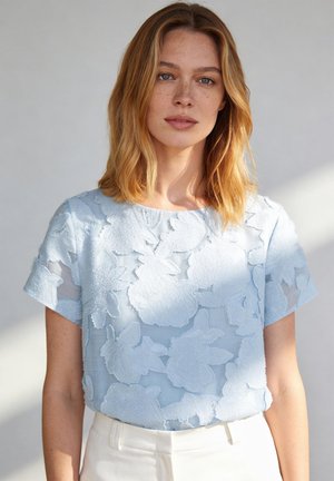 Femme aux cheveux blonds mi-longs, portant un chemisier à manches courtes bleu clair à texture florale et un pantalon blanc, sur un fond uni.