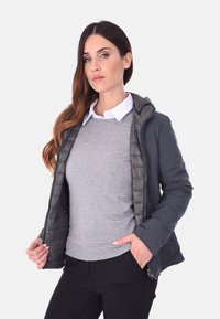 Giacca a vento grigia con cappuccio, dotata di una superficie esterna liscia, texture trapuntata e cerniere. Indossata sopra un maglione grigio chiaro e pantaloni neri.
