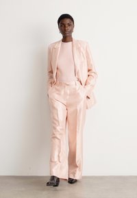 Woman stands against white wall wearing a pale pink satin suit with wide-leg trousers, matching sweater, and black pointed shoes.