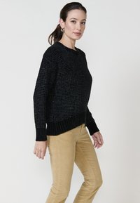 Črna pletena jopica z teksturiranim dizajnom in rebrastimi manšetami, v kombinaciji s svetlobrnimi korodnimi hlačami. Model stoji v profilu.