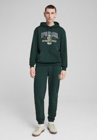 Felpa con cappuccio verde scuro e pantaloni jogging abbinati, dotati di un logo grafico con la scritta "PUMA Sports Legacy 1949." Tessuto di cotone con polsini e vita a coste.