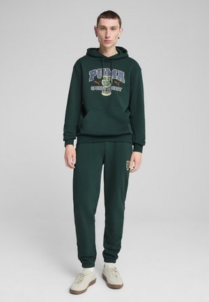 Felpa con cappuccio verde scuro e pantaloni jogging abbinati, dotati di un logo grafico con la scritta "PUMA Sports Legacy 1949." Tessuto di cotone con polsini e vita a coste.