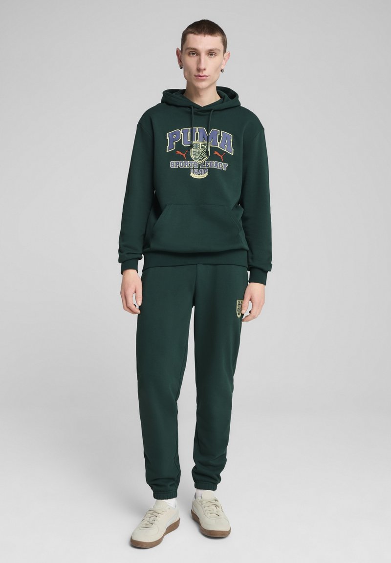 Felpa con cappuccio verde scuro e pantaloni jogging abbinati, dotati di un logo grafico con la scritta "PUMA Sports Legacy 1949." Tessuto di cotone con polsini e vita a coste.