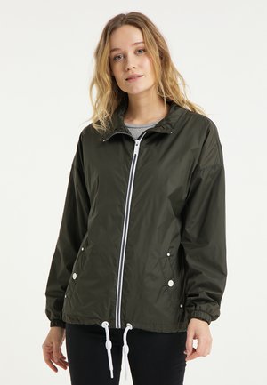 Jeune femme aux cheveux clairs portant une veste zippée vert foncé avec des cordons blancs et un pantalon noir, debout devant un fond uni.