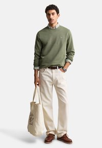 Homme debout portant un pull vert olive, une chemise rayée, un pantalon beige, des chaussures marron, tenant un sac fourre-tout beige avec du texte imprimé.