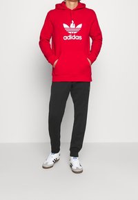 Červená mikina s kapucí s bílým logem Adidas a třemi pruhy; doplněna černými tepláky a bílými teniskami s černými detaily.