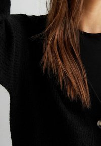Černý strukturovaný kardigan s knoflíkovou detailou, v popředí dlouhé rovné vlasy na neutrálním pozadí.