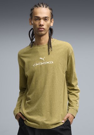 Jeune homme aux cheveux tressés portant un t-shirt Puma à manches longues vert olive et un pantalon noir, debout les mains dans les poches.