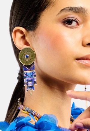 Femme portant de grandes boucles d'oreilles rondes en or et perles bleues ainsi qu'un collier assorti multicolore en perles, avec un haut bleu à volants.
