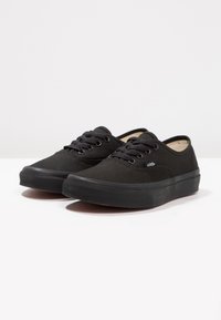 Zapatillas de lona negras con puntera redonda, suela plana de goma y diseño de cordones. Presenta una etiqueta del logo de Vans en el costado y parte superior texturizada.
