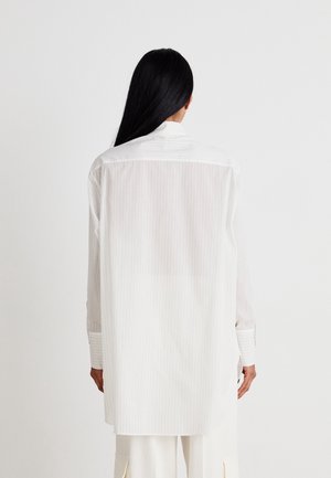 Osoba z długimi czarnymi włosami, ubrana w luźną, białą koszulę w cienkie prążki z długim rękawem i białe spodnie, zwrócona tyłem na jednolitym tle.