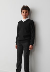 Črni pulover z V-izrezom iz mehke tkanine, kombiniran s črnimi ohlapno prilegajočimi hlačami. Obleka vključuje belo srajco z ovratnikom pod njim.