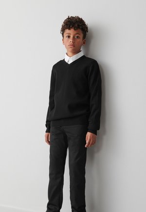 Fekete V-nyakú pulóver puha anyagból, fekete, slim fit nadrággal párosítva. A ruha alatt fehér galléros ing látható.