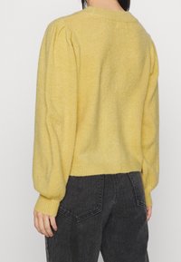 Žena v měkkém žlutém pleteném svetru s nafouknutými dlouhými rukávy a tmavých džínách s vysokým pasem, zobrazená zezadu.