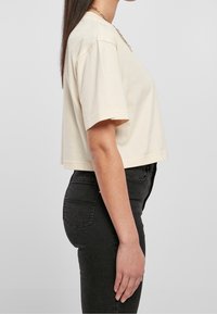 Donna con capelli scuri e lisci, che indossa un crop top beige, jeans neri a vita alta e una collana d'oro, in piedi di profilo.