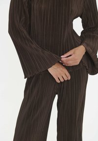 Persona che indossa una maglia a maniche larghe a coste marrone scuro e pantaloni larghi abbinati, mentre aggiusta il fondo della maglia.