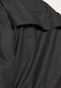 Detailní záběr na černou voděodolnou látku bundy, ukazující hladkou texturu a detailem kápě na zádech.