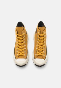 High-top sneakers with a mustard-yellow canvas upper, black accents, white rubber toe cap, and black textured sole with white midsole.