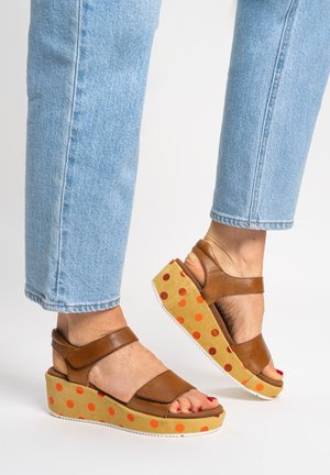 Füße, die braune Leder-Plateau-Sandalen mit orangefarbenen Punkten auf der Sohle tragen, kombiniert mit hellblauen Jeans.