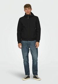 Black zip-up jacket with a hood, featuring a smooth fabric and two front pockets. Paired with blue denim jeans and black sneakers.