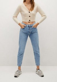 Cárdigan corto de canalé beige con tres botones, combinado con vaqueros de tiro alto en azul claro. Pies con zapatillas deportivas grises con cordones blancos.