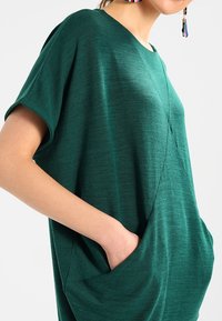 Tunica verde a maniche corte realizzata in tessuto morbido. Presenta una vestibilità rilassata, scollatura rotonda e tasche laterali. Dettaglio della superficie strutturata visibile.