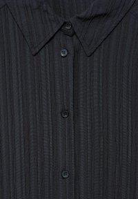 Camicia nera con texture e righe verticali, dotata di colletto e quattro bottoni. Il tessuto appare leggero e leggermente stropicciato.