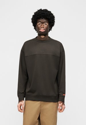 Άτομο με λεύκη και σγουρά μαλλιά, φοράει σκούρο πουκάμισο μακρυμάνικο και μπεζ παντελόνι, στέκεται μπροστά σε απλό ανοιχτό φόντο.