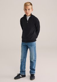 Zēns stāv ar rokām aiz muguras, ģērbies melnā ceturtdaļas rāvējslēdzēja džemperī, zilos džinsos un melnās kedās uz neitrālas fona.