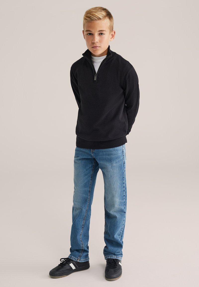 Zēns stāv ar rokām aiz muguras, ģērbies melnā ceturtdaļas rāvējslēdzēja džemperī, zilos džinsos un melnās kedās uz neitrālas fona.