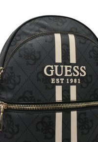 Schwarzer Kunstleder-Rucksack mit Canvas-Struktur, ausgestattet mit weißen vertikalen Streifen, dem Logo "GUESS EST 1981" und goldenen Reißverschlussteilen.