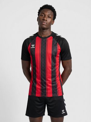Young man standing with hands behind back wearing red and black striped sports shirt and black shorts on white background.
